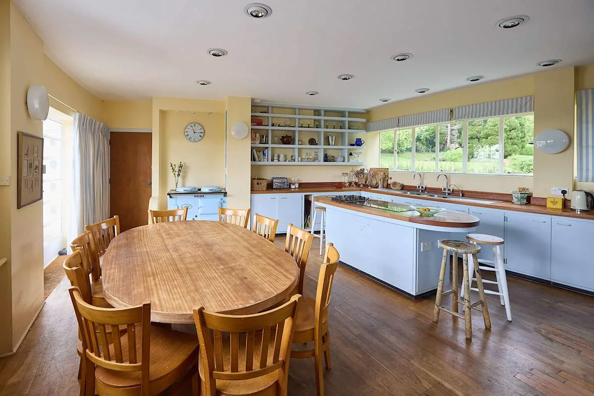 Una cocina luminosa con zona de comedor, una mesa larga de madera y una isla central.