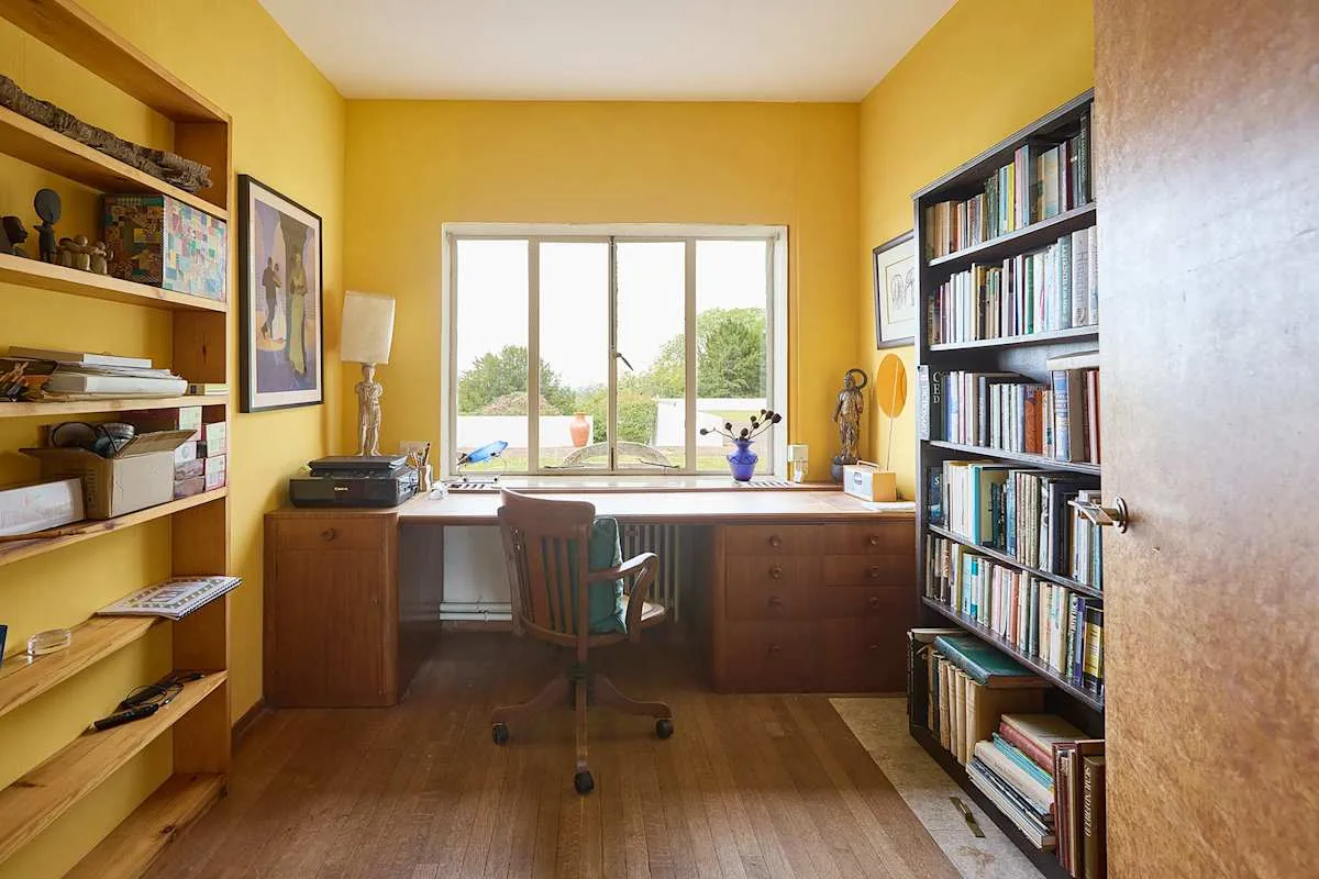 Un despacho en casa con un escritorio frente a una ventana grande y estanterías.