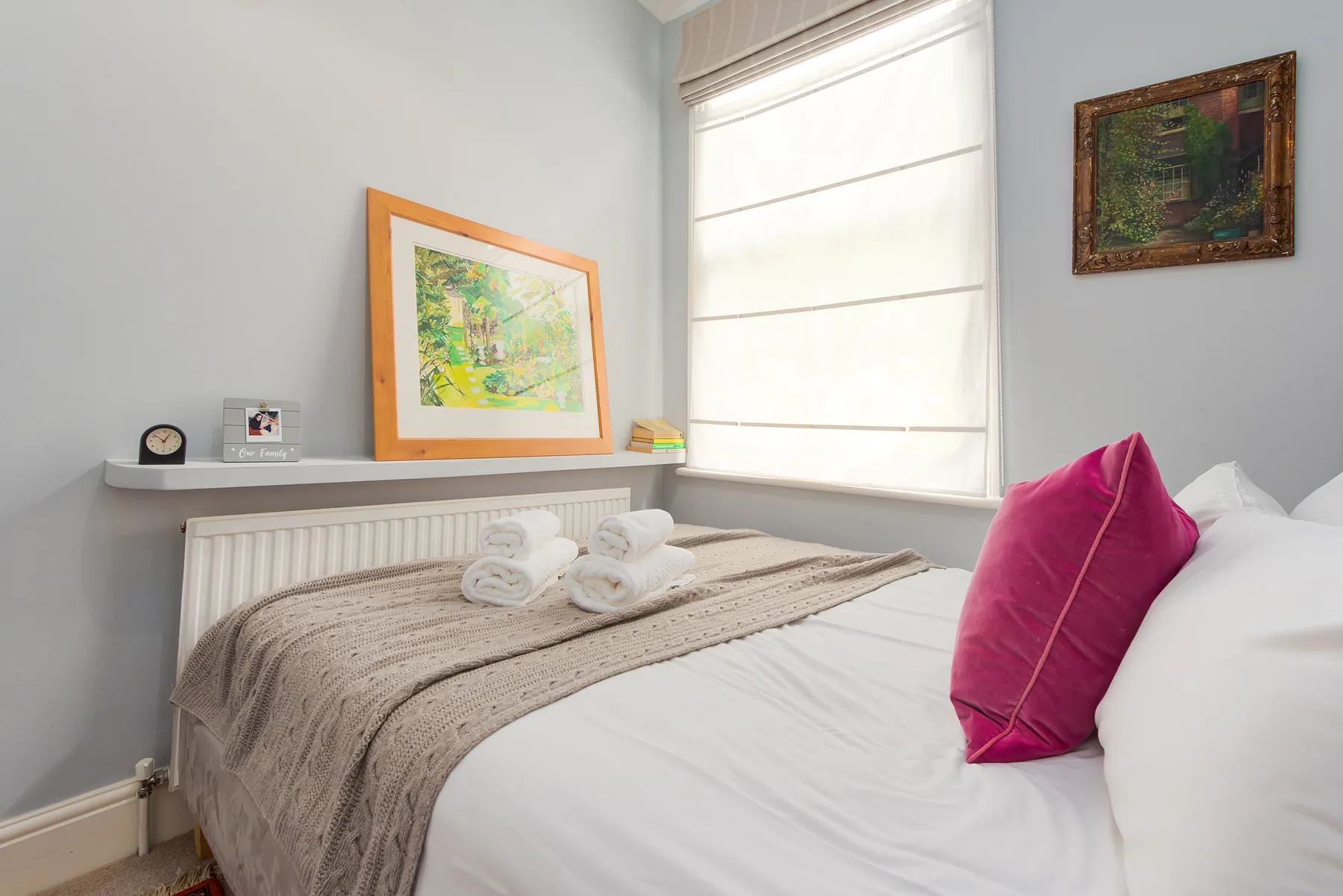 A bedroom with a neatly made bed, pink cushion, and framed artwork.