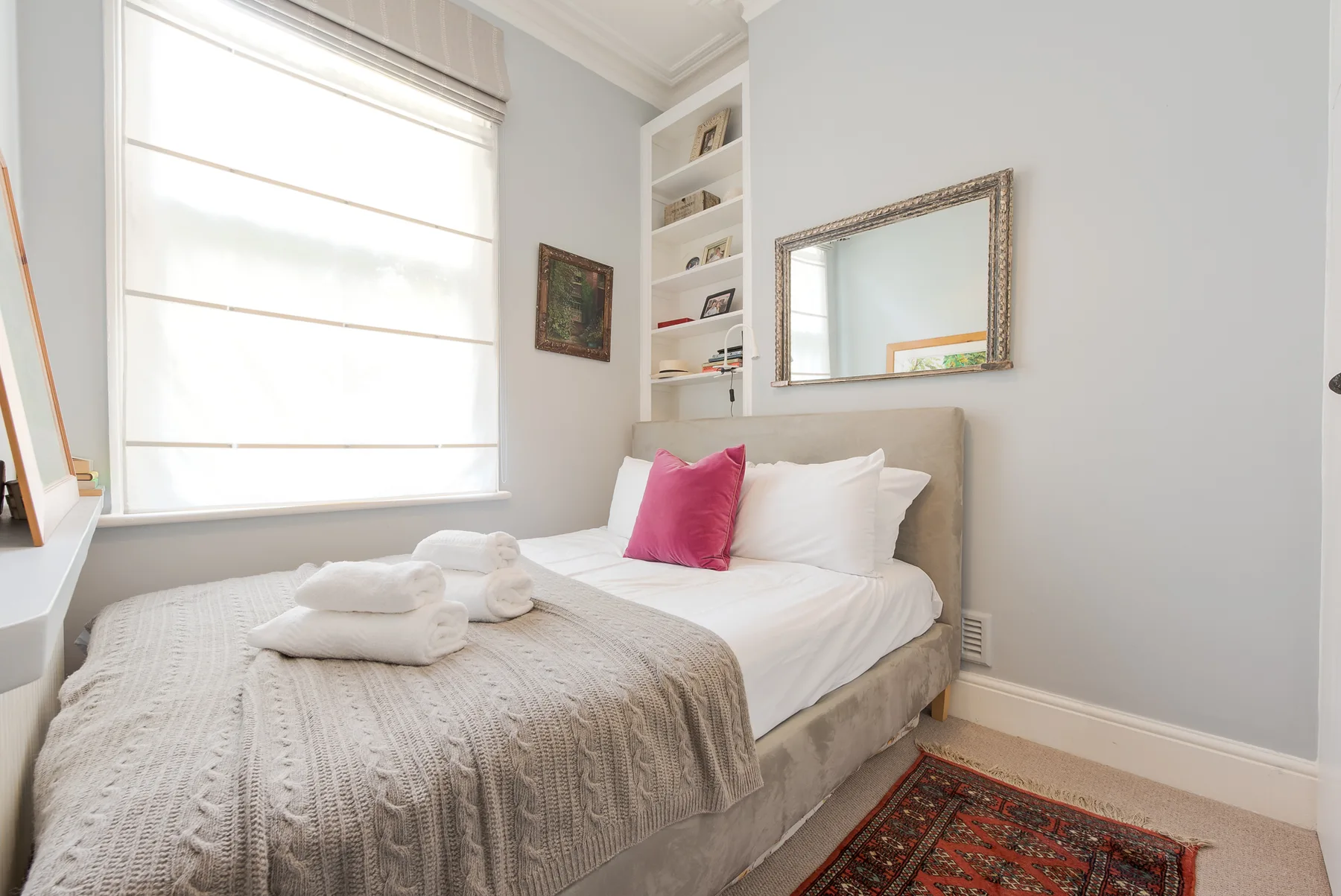 A bright bedroom with white bedding, a pink accent pillow, and built-in shelves.