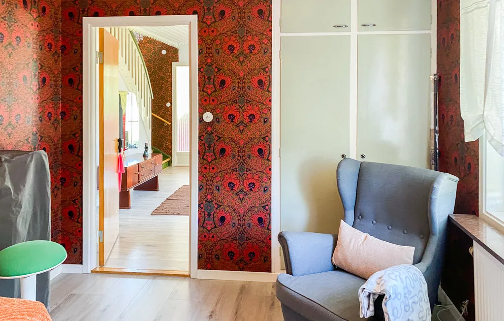 A sitting area with an upholstered chair beside a window and patterned wallpaper.