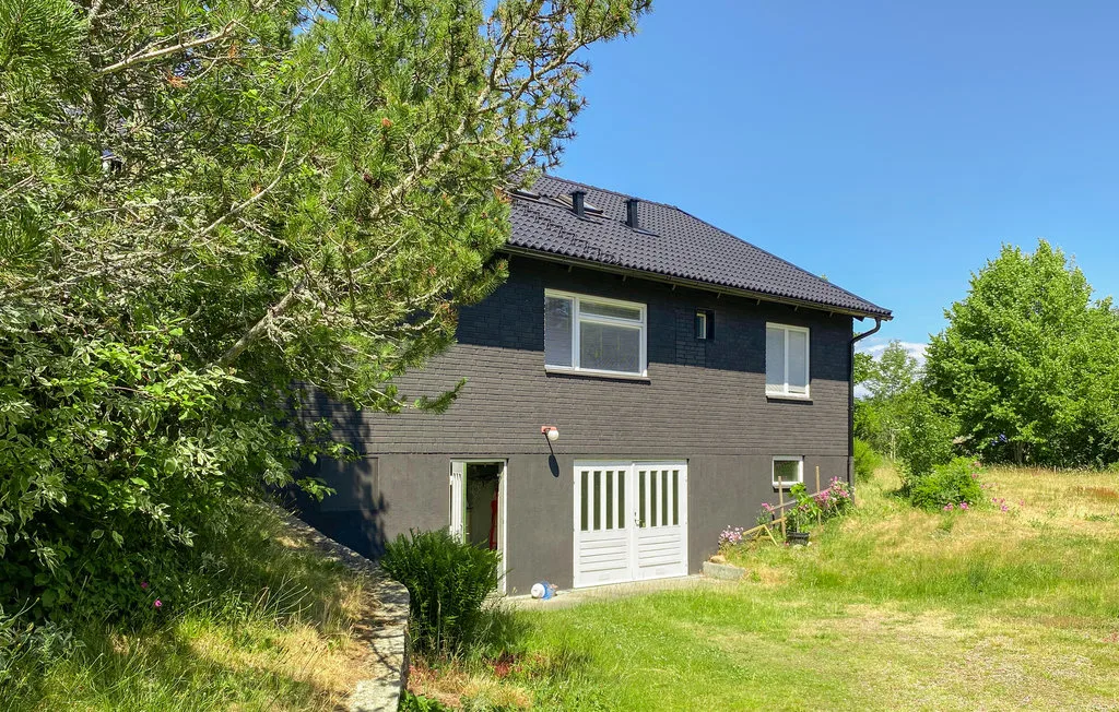 A dark two-story house stands beside a grassy yard and leafy trees.