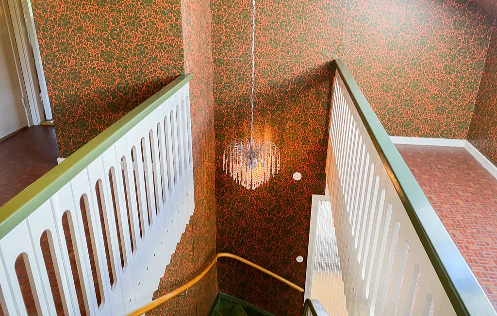 A staircase descends between white railings beside patterned red walls and a hanging light.