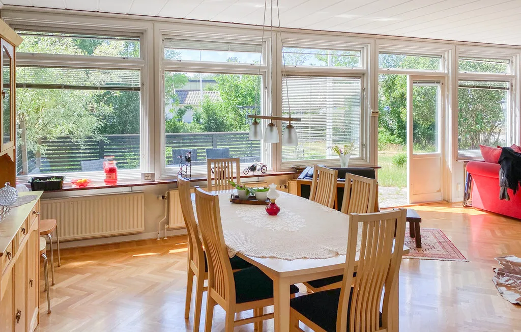 A bright dining area has a long table beside large windows overlooking greenery.
