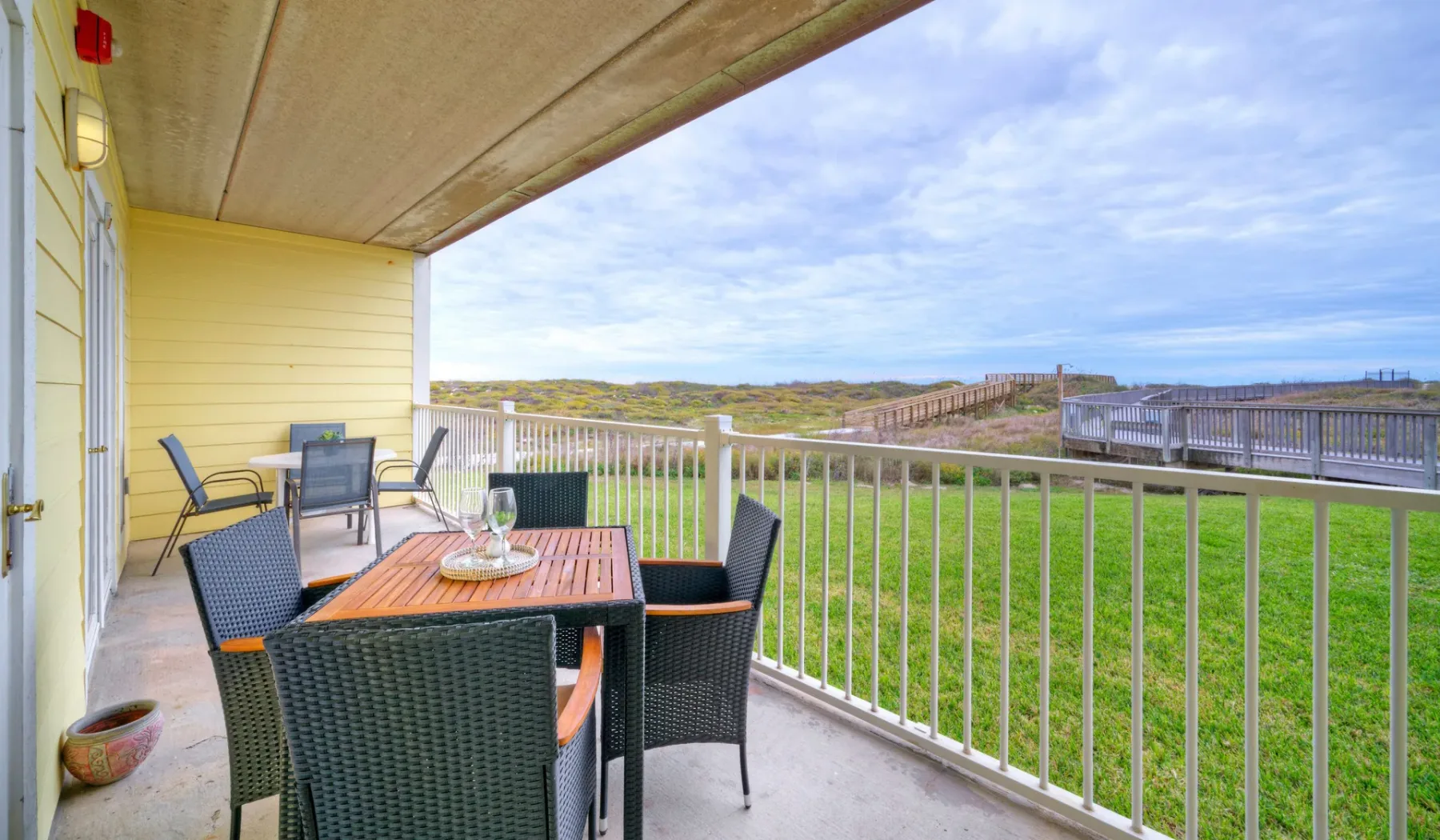 Un patio cubierto tiene una mesa de comedor junto a una barandilla blanca, con vista a una zona de césped.