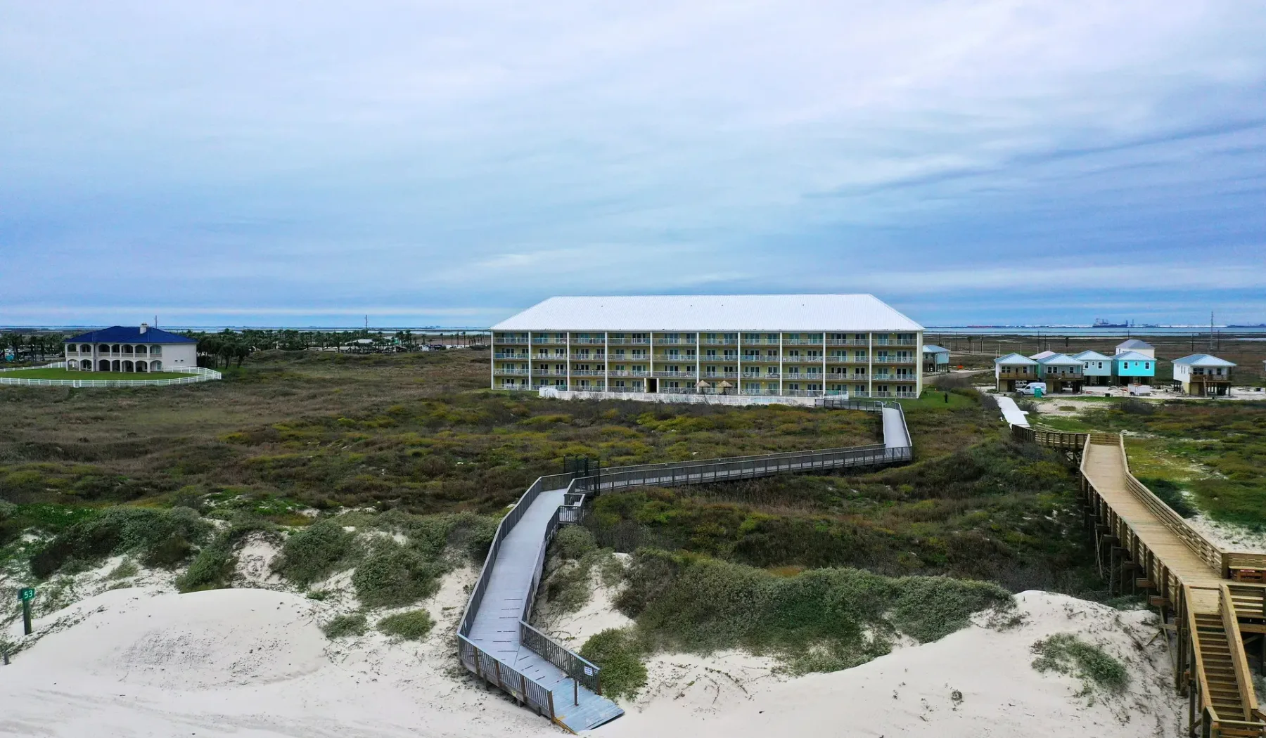 Paisaje costero con dunas de arena, pasarelas elevadas y edificios a lo lejos.