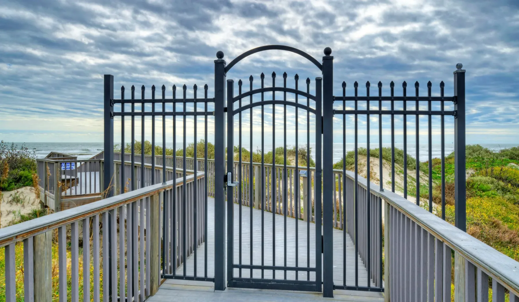 Una pasarela elevada atraviesa una puerta metálica negra hacia la playa.