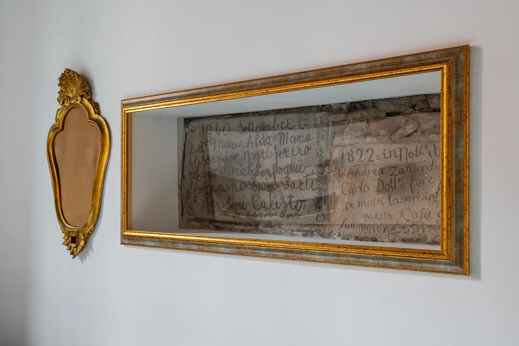Un documento manuscrito enmarcado y un pequeño espejo decorativo cuelgan de una pared clara.