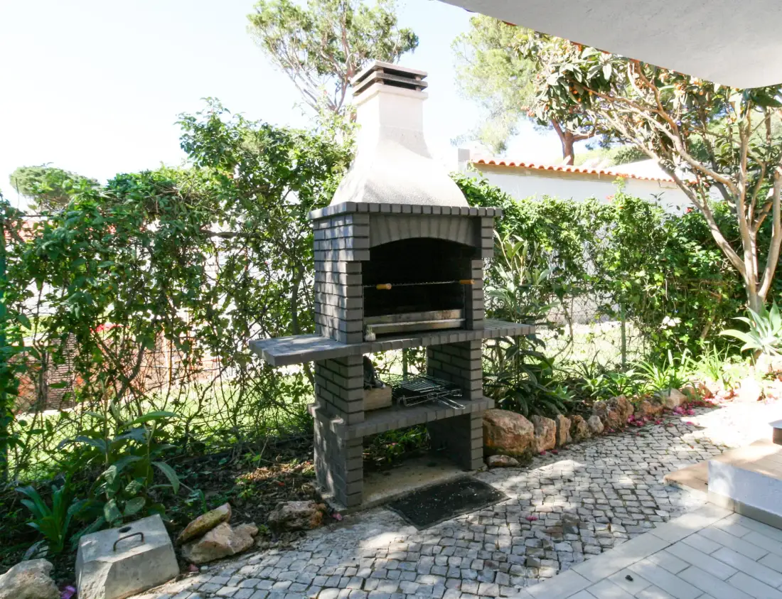 Una zona de barbacoa al aire libre junto a un sendero empedrado y un jardín frondoso.