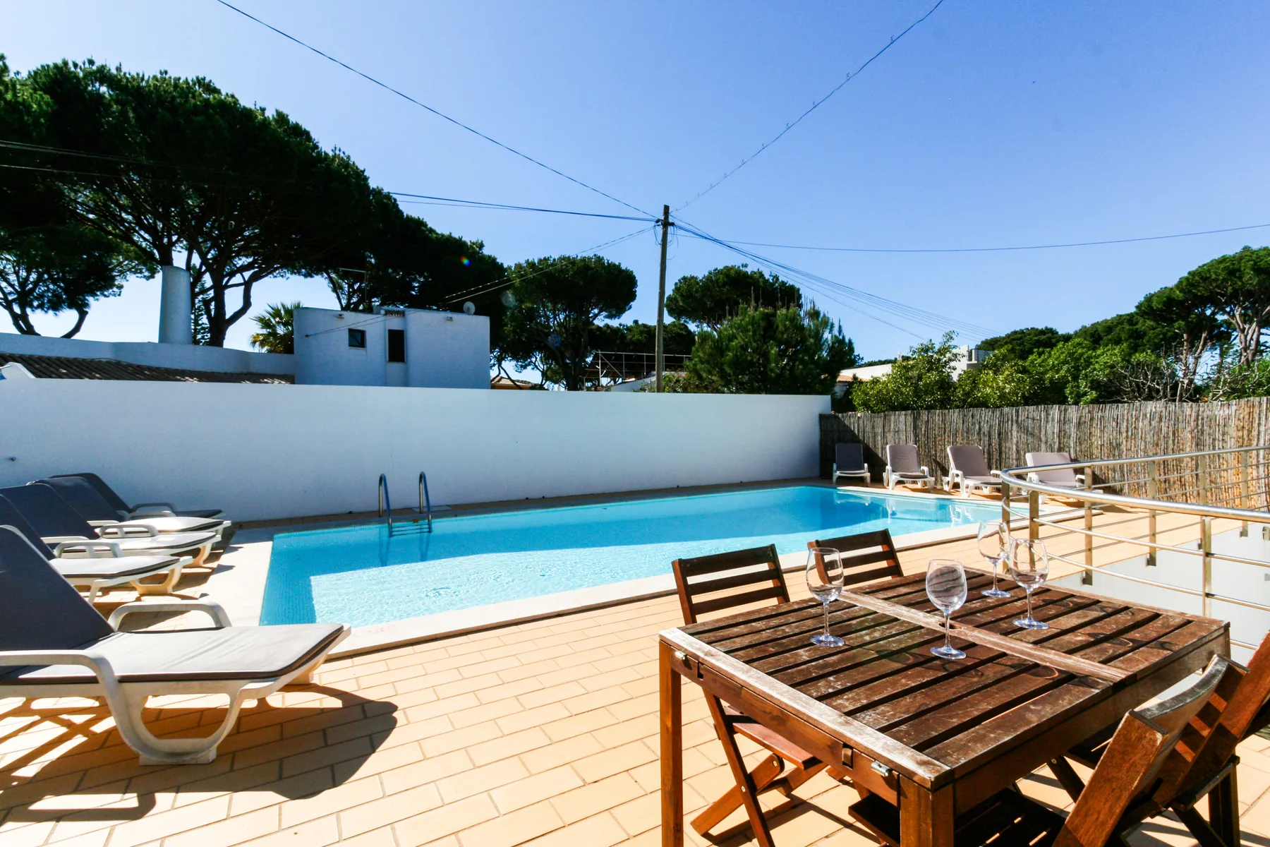 Una piscina al aire libre junto a un patio embaldosado con asientos y una mesa de madera.