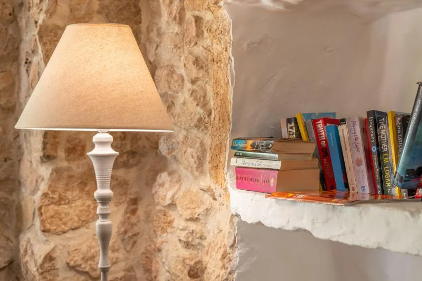 A table lamp stands beside books on a built-in shelf against a stone wall.