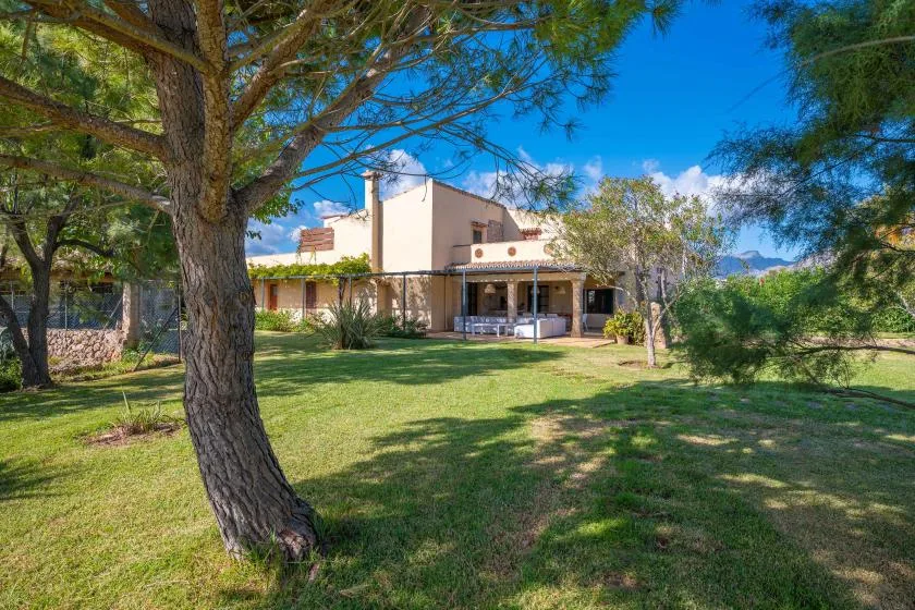 A house stands beyond a green lawn beneath mature trees.