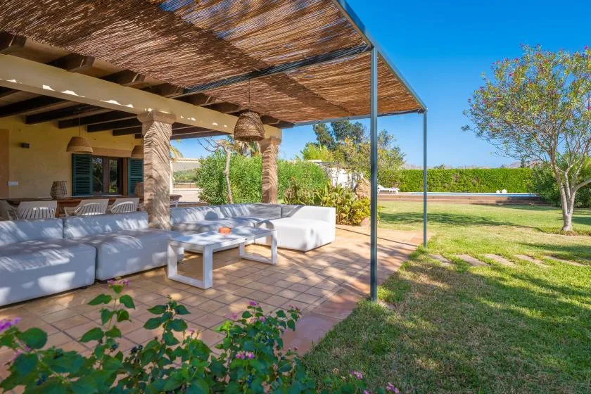 A shaded outdoor patio has seating and a table beside a grassy yard.