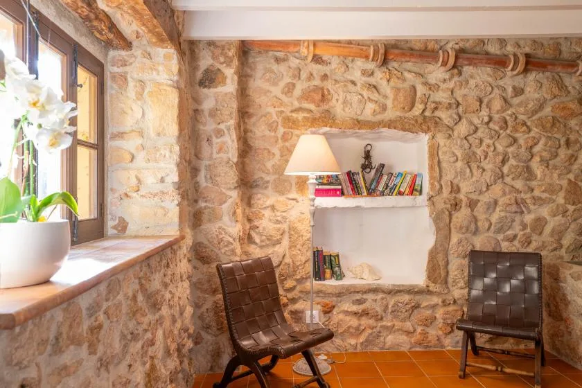 A stone-walled room with a recessed bookshelf, lamp, and seating.