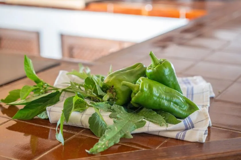 Green peppers and leafy herbs rest on a cloth-covered table.