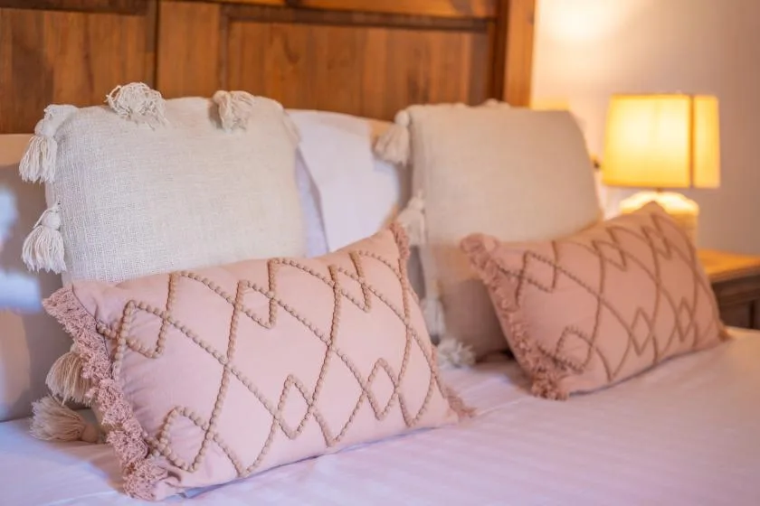 A bed with decorative pillows sits beneath a wooden wall, beside a lit bedside lamp.