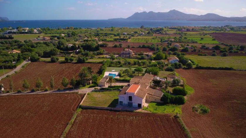 An aerial view of a farmhouse amid cultivated fields, with the sea and mountains beyond.
