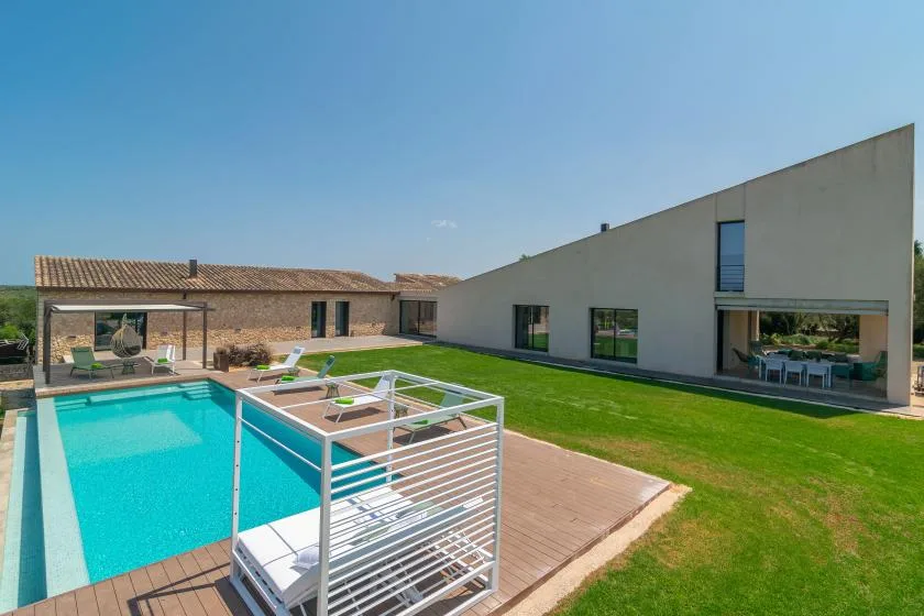A modern house opens onto a turquoise swimming pool and a green lawn.