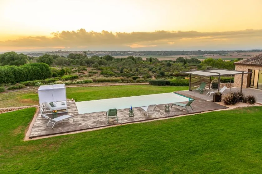 An outdoor swimming pool and patio overlook green fields beneath a sunset sky.