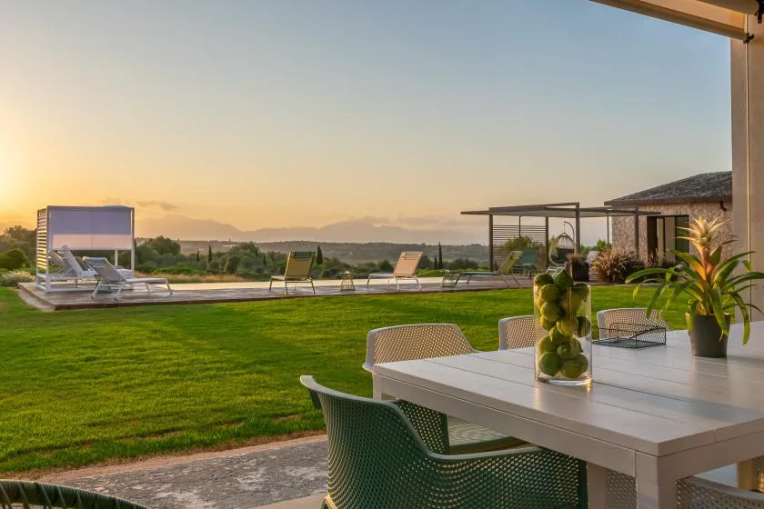 An outdoor dining area overlooks a lawn, pool, and distant hills at sunset.