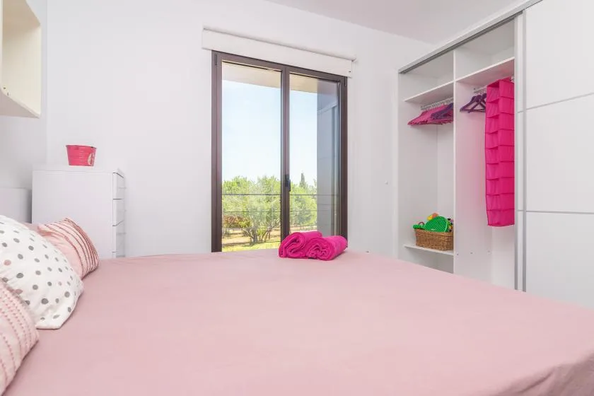 A bedroom with a pink-covered bed beside an open wardrobe and glass doors.