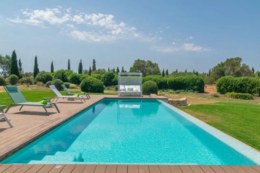 An outdoor swimming pool bordered by a wooden deck, lounge seating, and green landscaping.