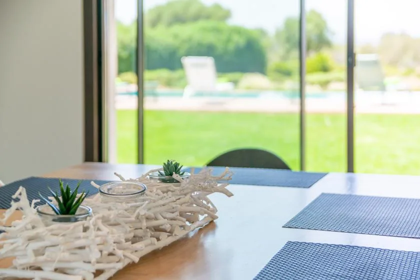 A dining table with placemats sits beside large windows overlooking a garden.