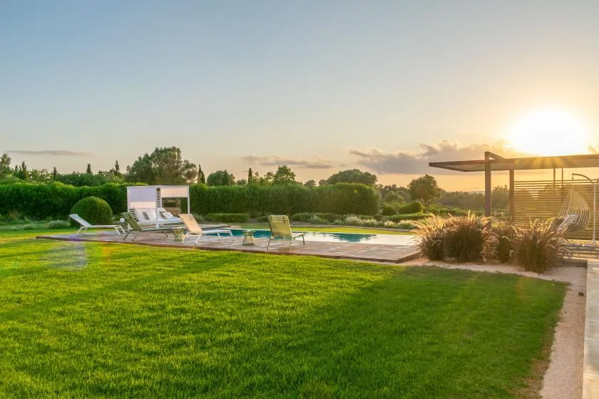 An outdoor swimming pool beside a lawn and lounge seating at sunset.
