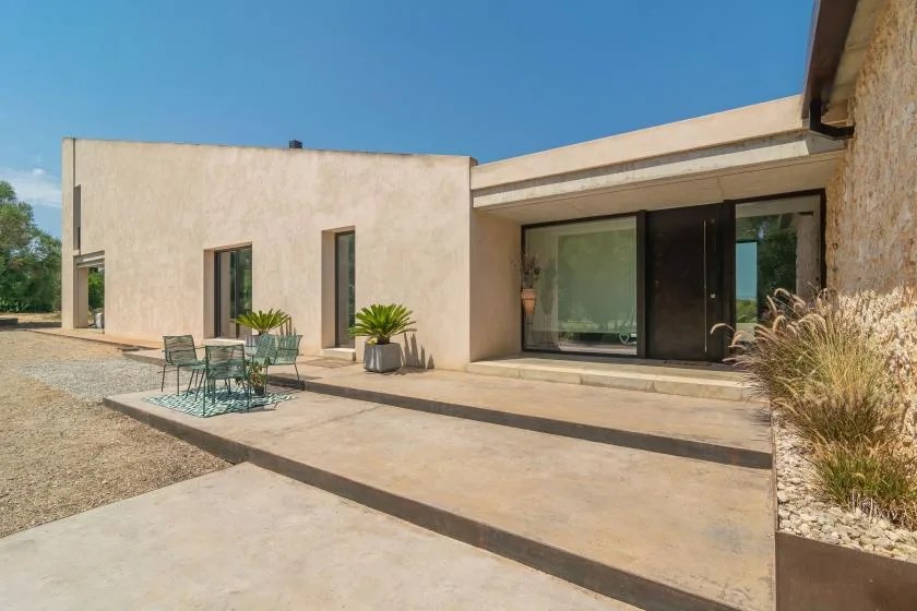 A modern house opens onto a broad paved patio with outdoor seating and sliding glass doors.