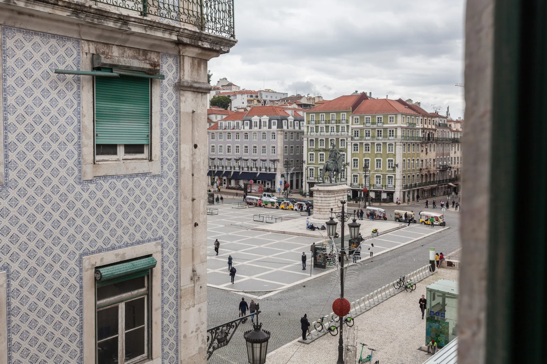 Vista de una plaza desde una ventana, con azulejos estampados en la fachada cercana.