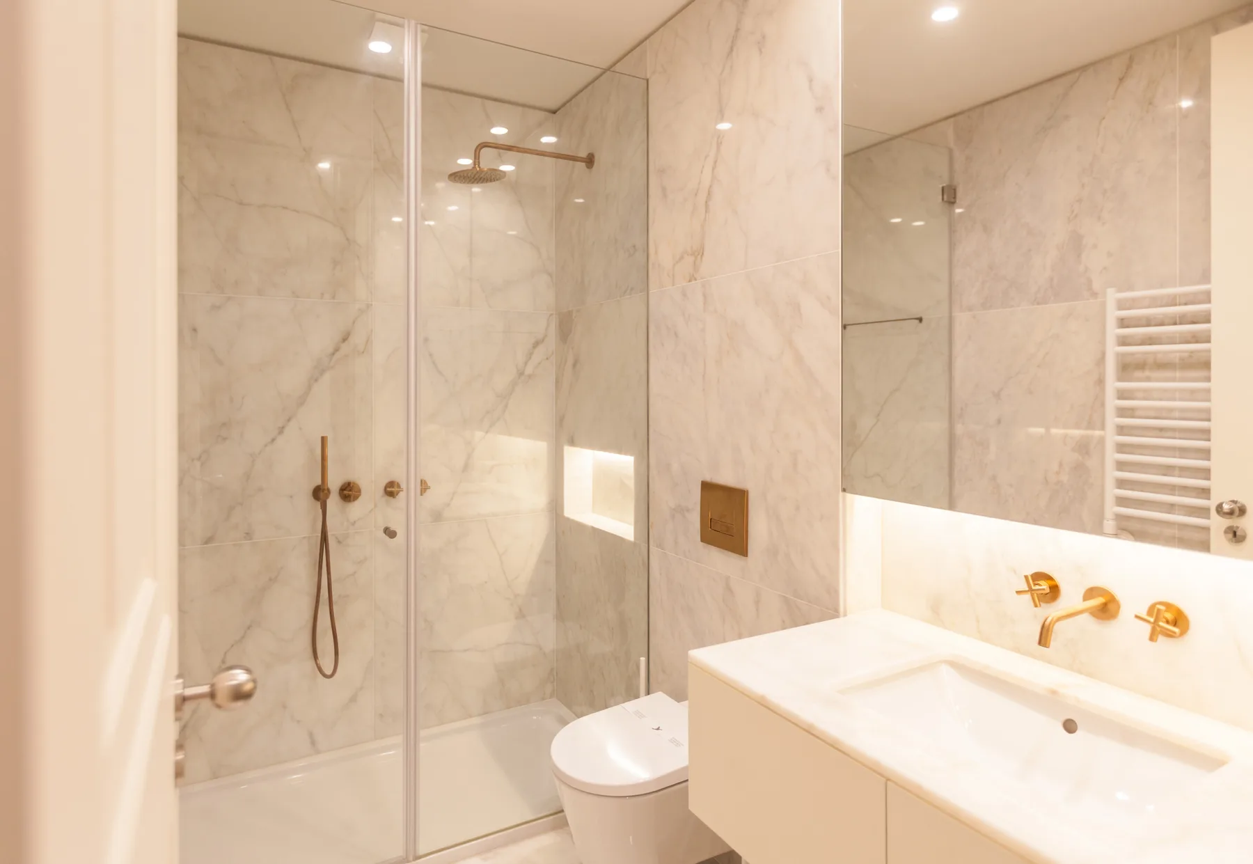 Un baño luminoso de mármol con ducha de vidrio, inodoro y lavabo.