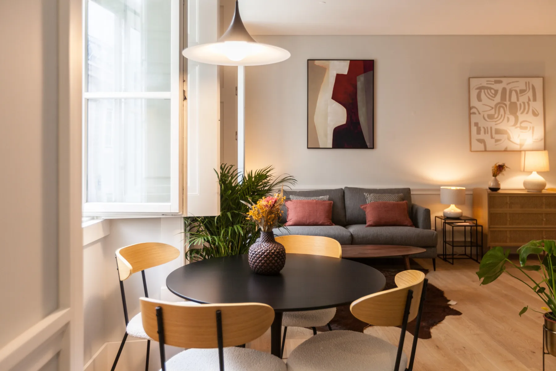Una sala de estar con un sofá gris, una mesa redonda y ventanas grandes.
