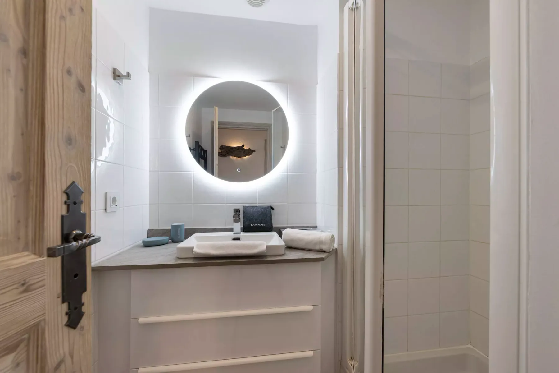 Un lavabo de baño con espejo redondo iluminado y una ducha al lado.