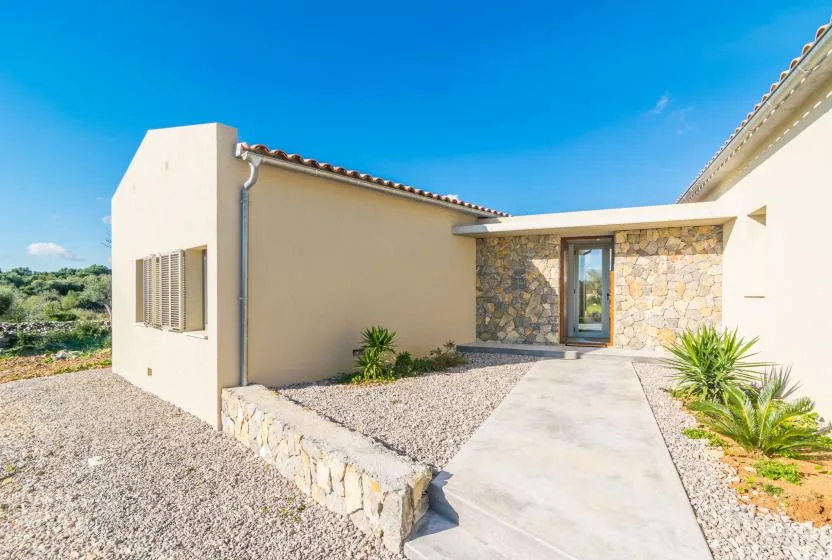 A sunlit house entrance has a stone facade and a paved path through gravel landscaping.