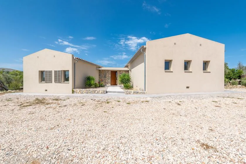 A low, light-colored building stands beyond a broad gravel area under a blue sky.
