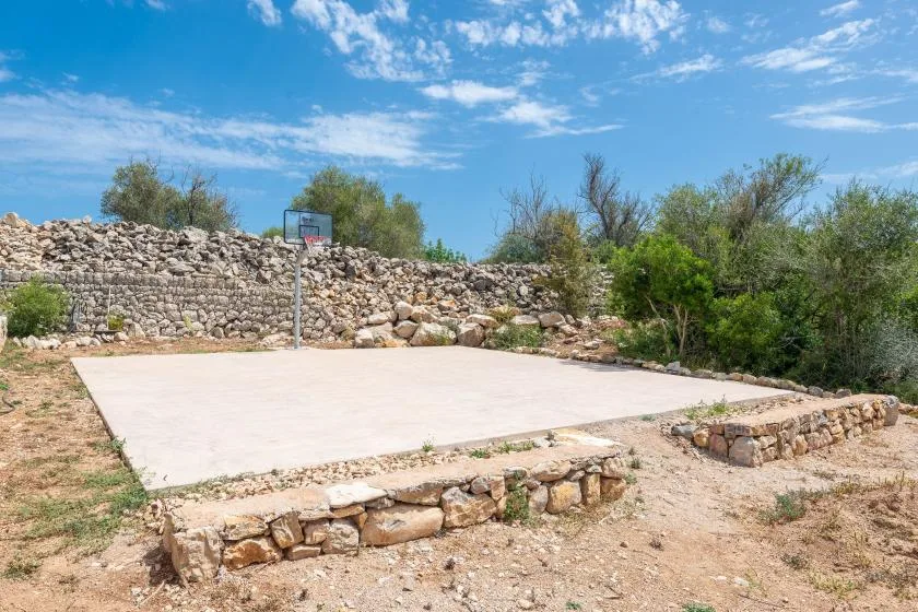 An outdoor basketball court sits beside a low stone wall and sparse shrubs.