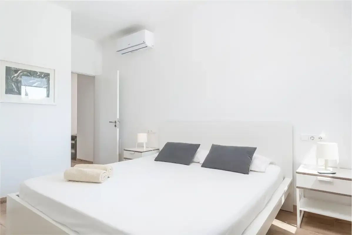 A bright bedroom with a bed, bedside tables, and a wall-mounted air conditioner.