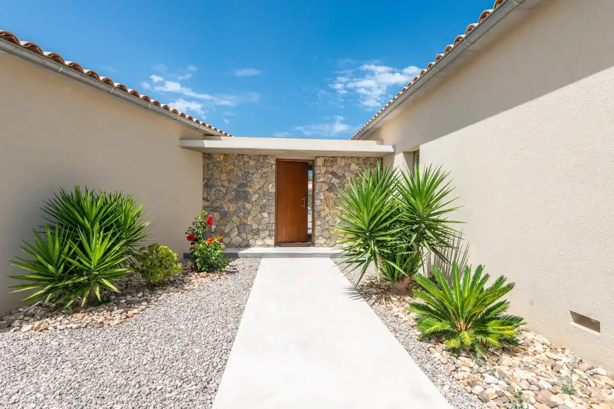 A paved walkway leads to a wooden entrance door between gravel garden beds.