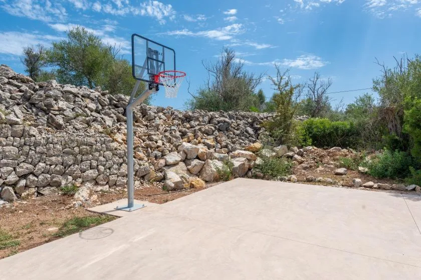 An outdoor basketball court sits beside a rocky wall and low shrubs.