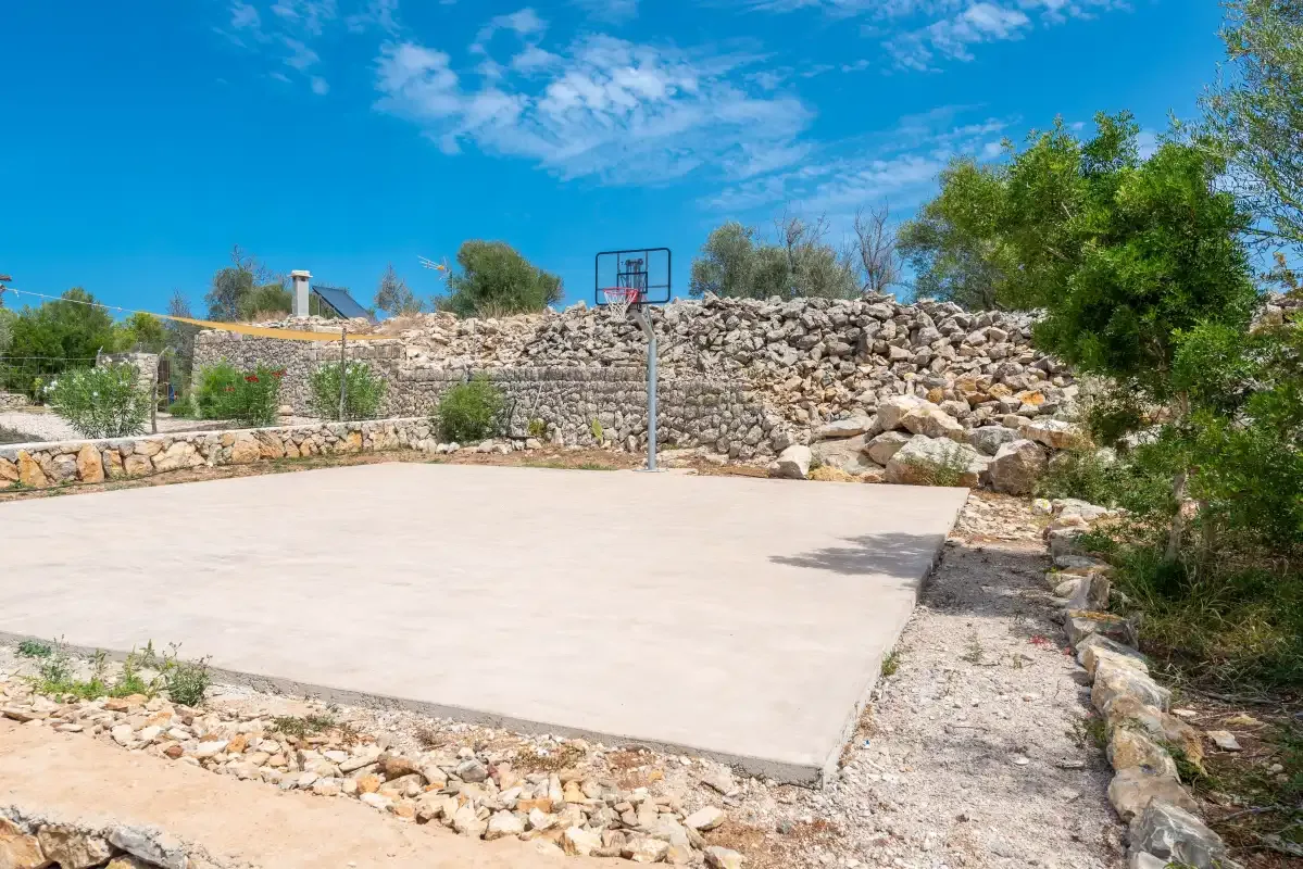 An outdoor basketball court sits beside a low stone wall and greenery.