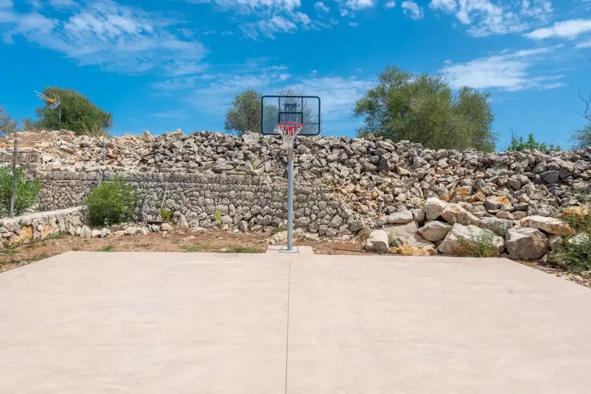 An outdoor basketball court stands in front of a stone wall under a blue sky.