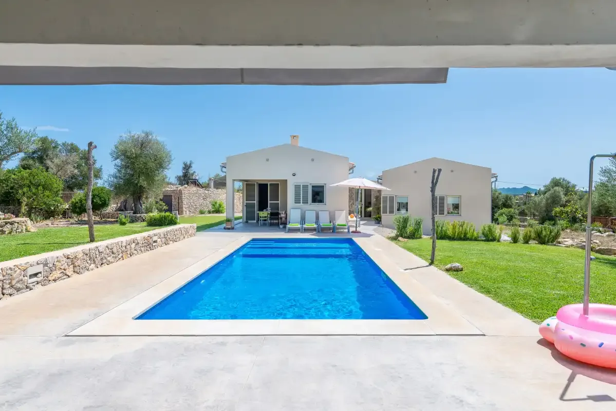 A rectangular swimming pool sits in front of a modern house, surrounded by lawn and paved areas.