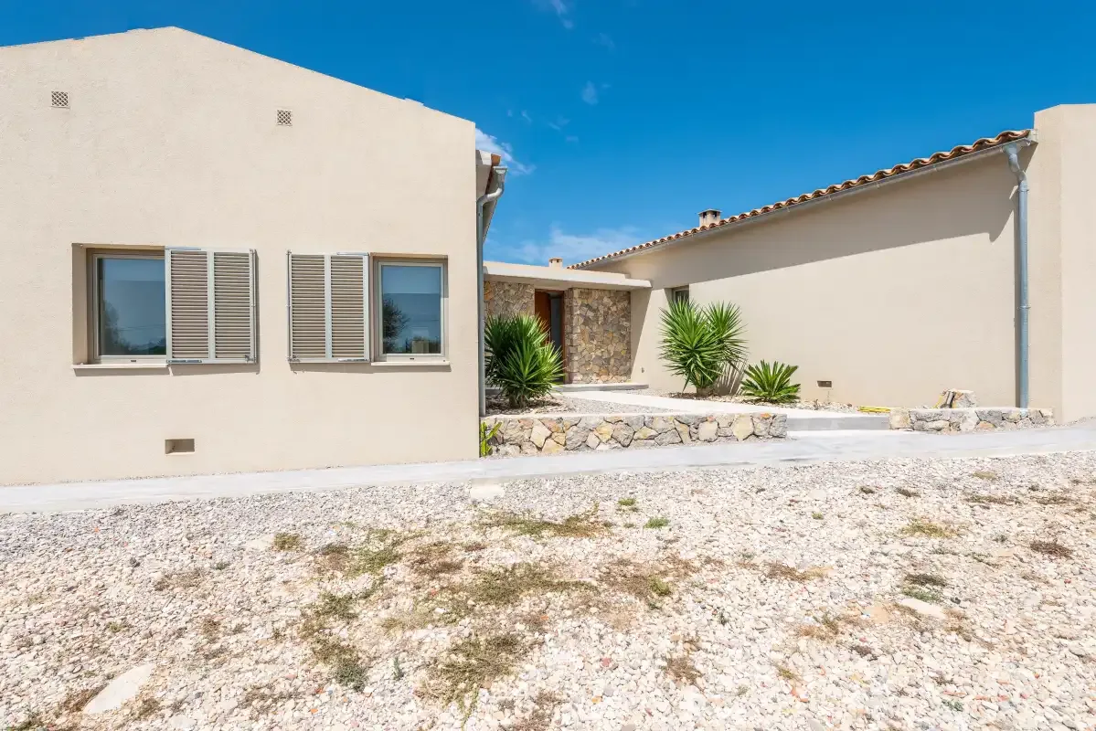 A pale stucco house faces a gravel courtyard with several plants.