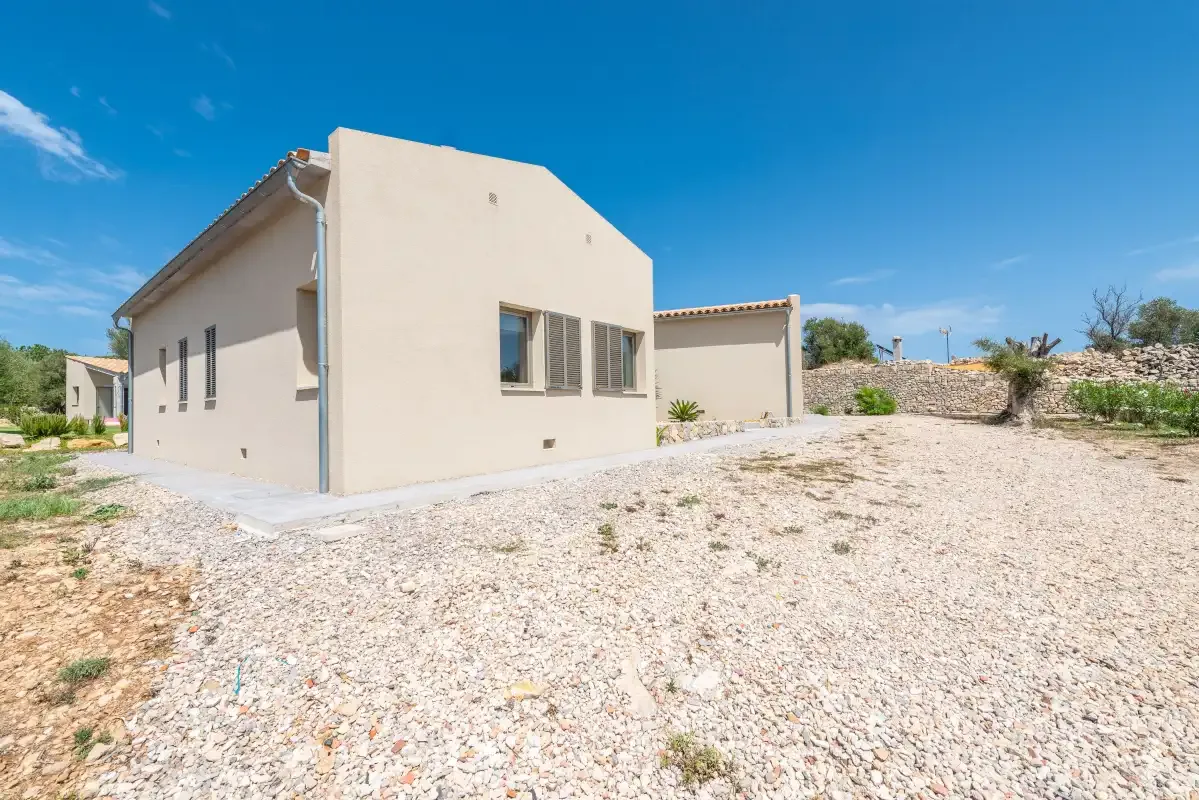 A pale stucco house stands beside a broad gravel yard under a clear blue sky.