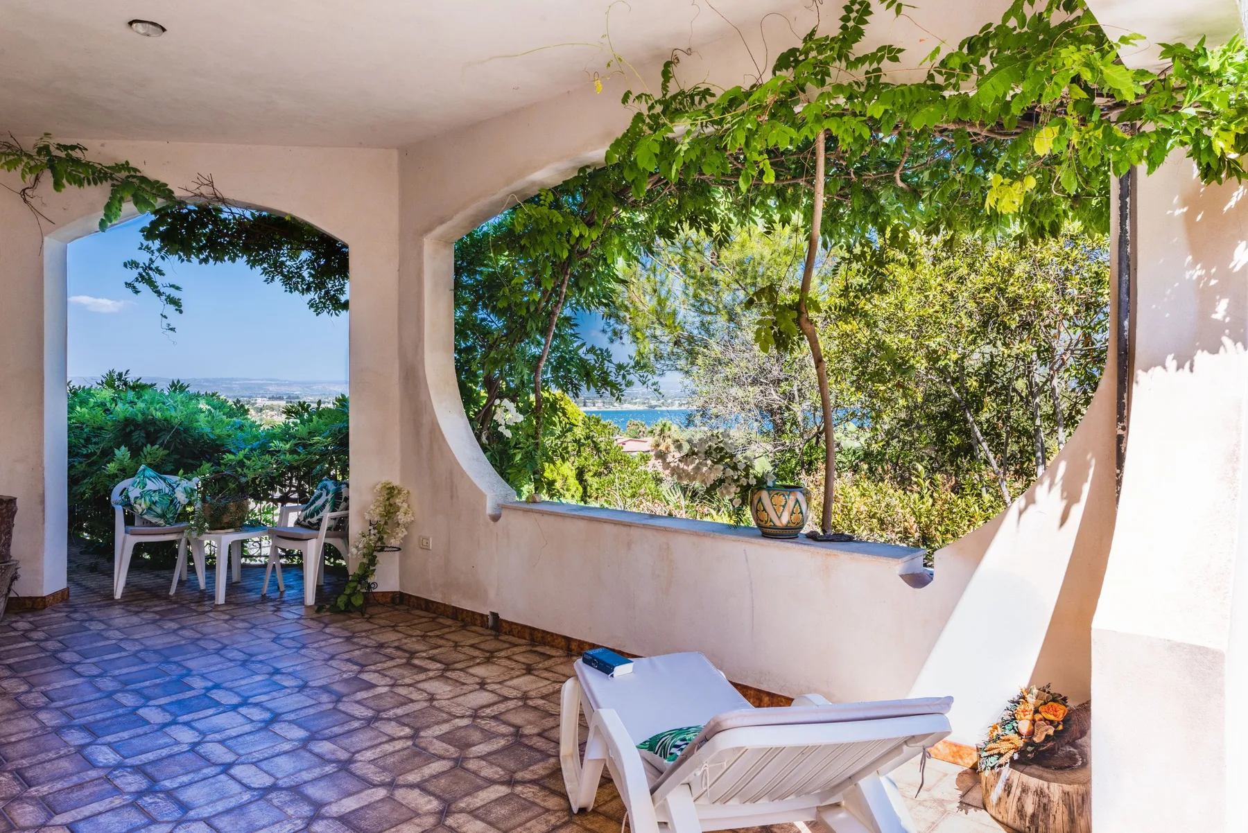 A covered patio with tiled flooring, seating, and leafy views toward the water.