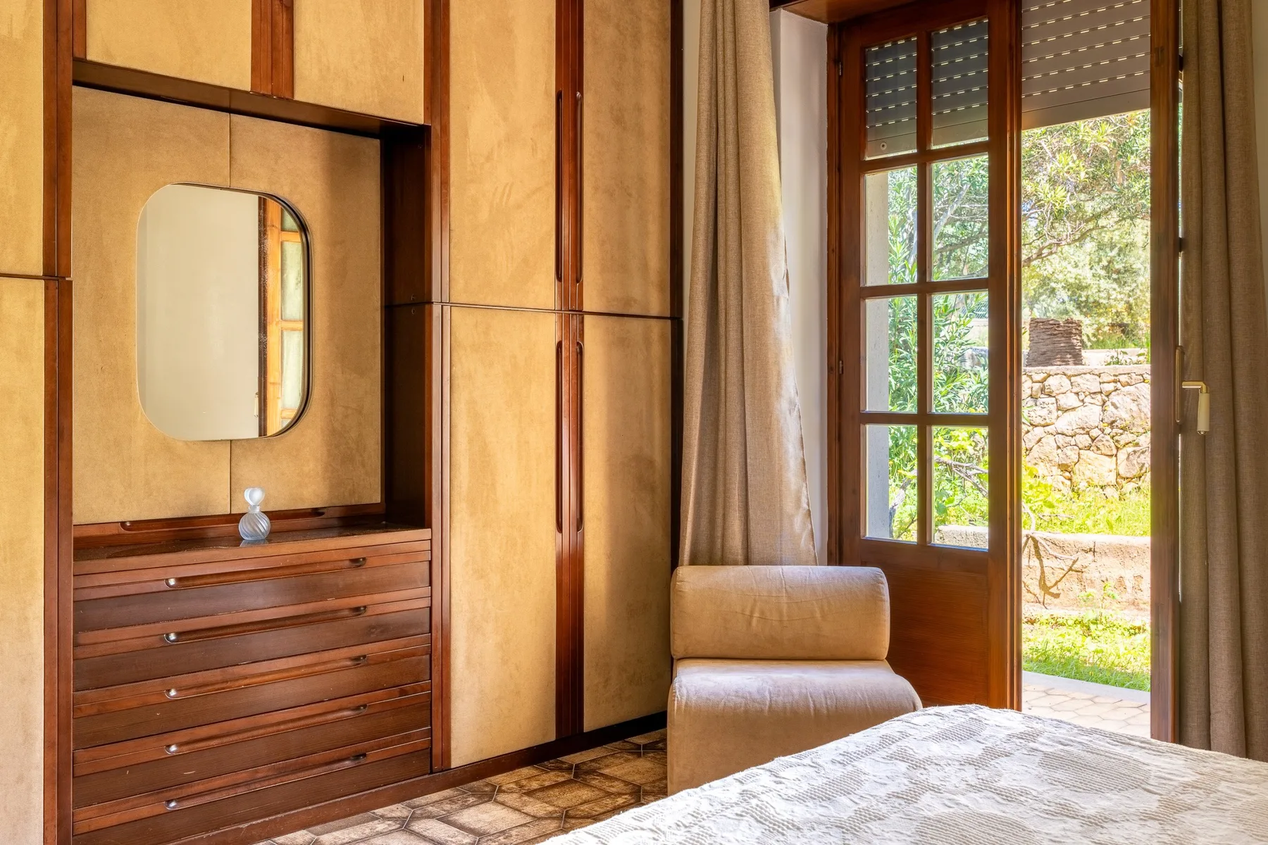 A bedroom with built-in wooden storage, a chair, and a window overlooking greenery.
