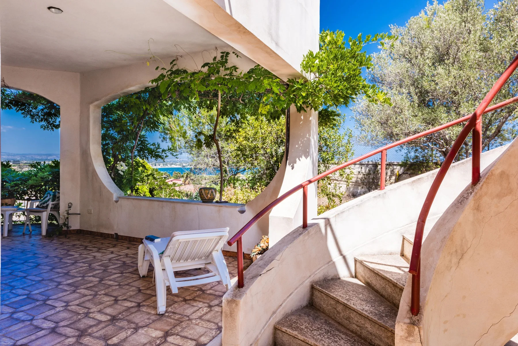 A shaded outdoor patio with seating overlooks trees and a blue coastline.