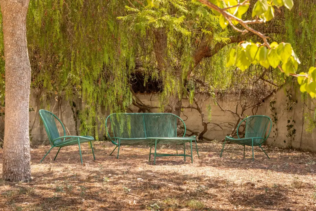 A shaded outdoor seating area with green metal chairs and a bench beneath leafy trees.