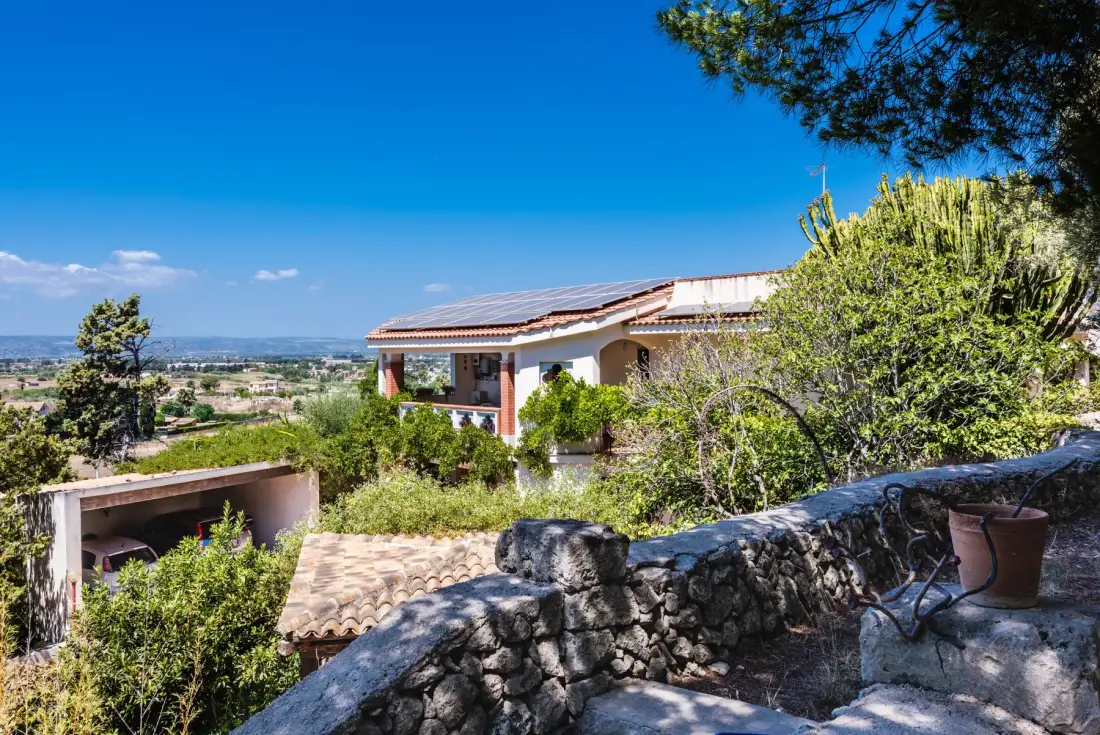 A house sits among trees and shrubs behind a stone wall on a sunny day.