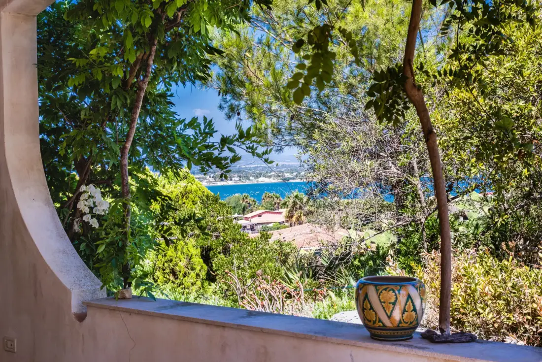 A leafy terrace overlooks the blue sea, with a decorative pot on the ledge.